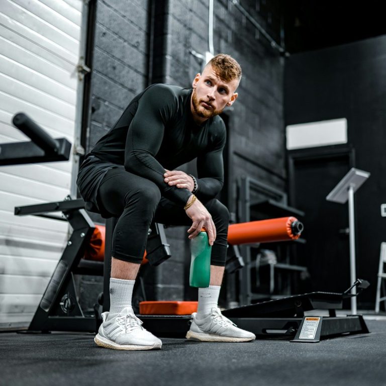 Een man in sportkleding zit op een fitnessbank en houdt een flesje water vast.